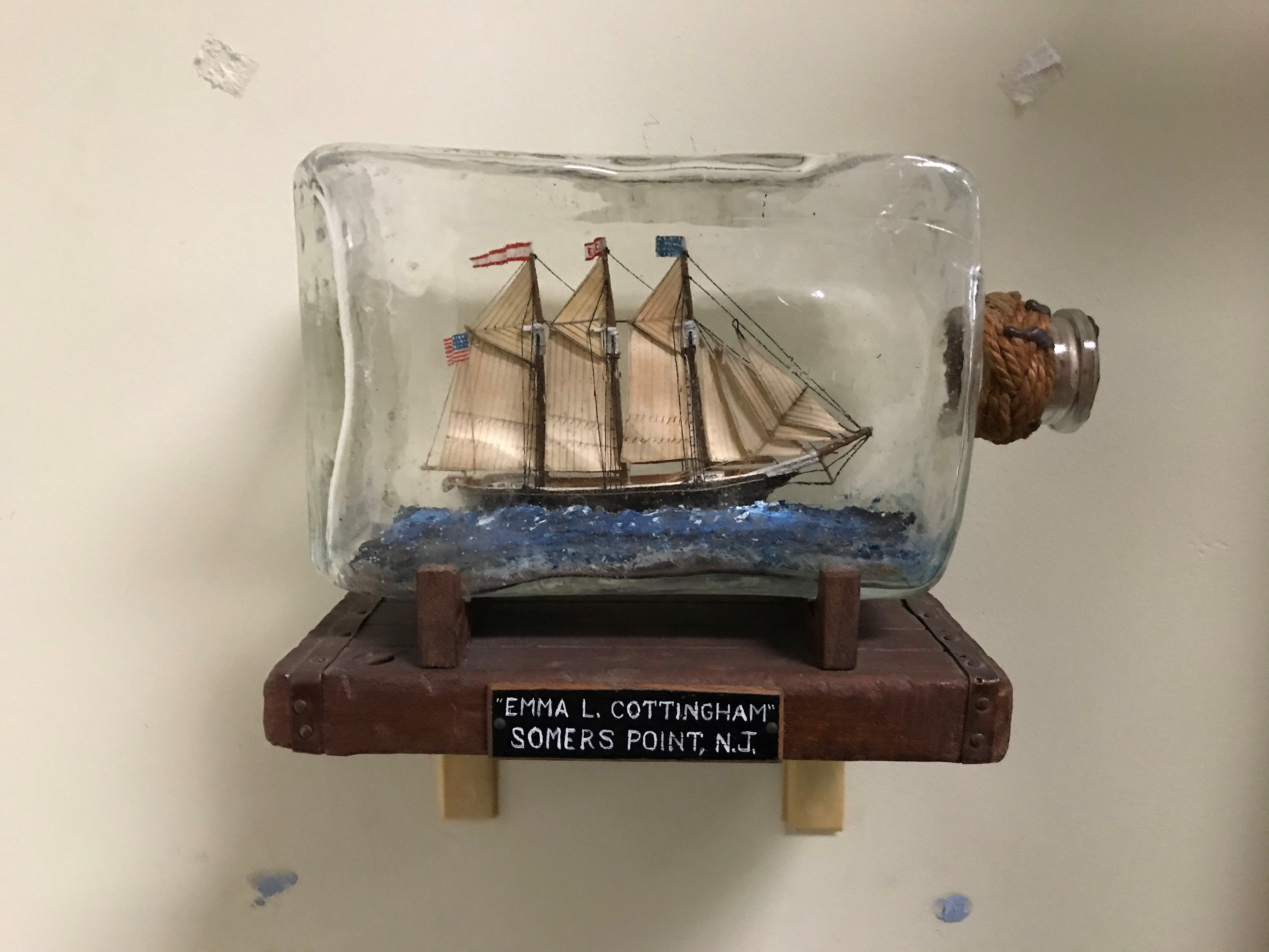small ship model inside a glass bottle, mounted on a shelf. Label indicates Emma L. Cottingham, Somers Point, NJ.