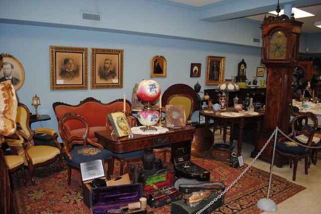 Image of a room arranged to look like an old salon, with armchairs, sofa, table, tall clock on a rug. Various items like photographs, musical instruments, and candlesticks add to the decor.