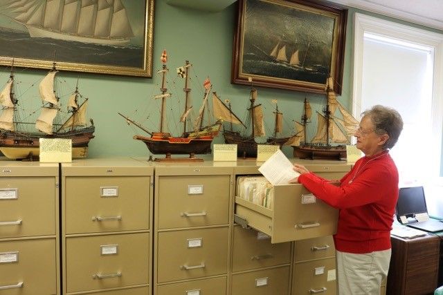 Woman looking at paper in filing drawer. Model ships on top of filing cabinet.
